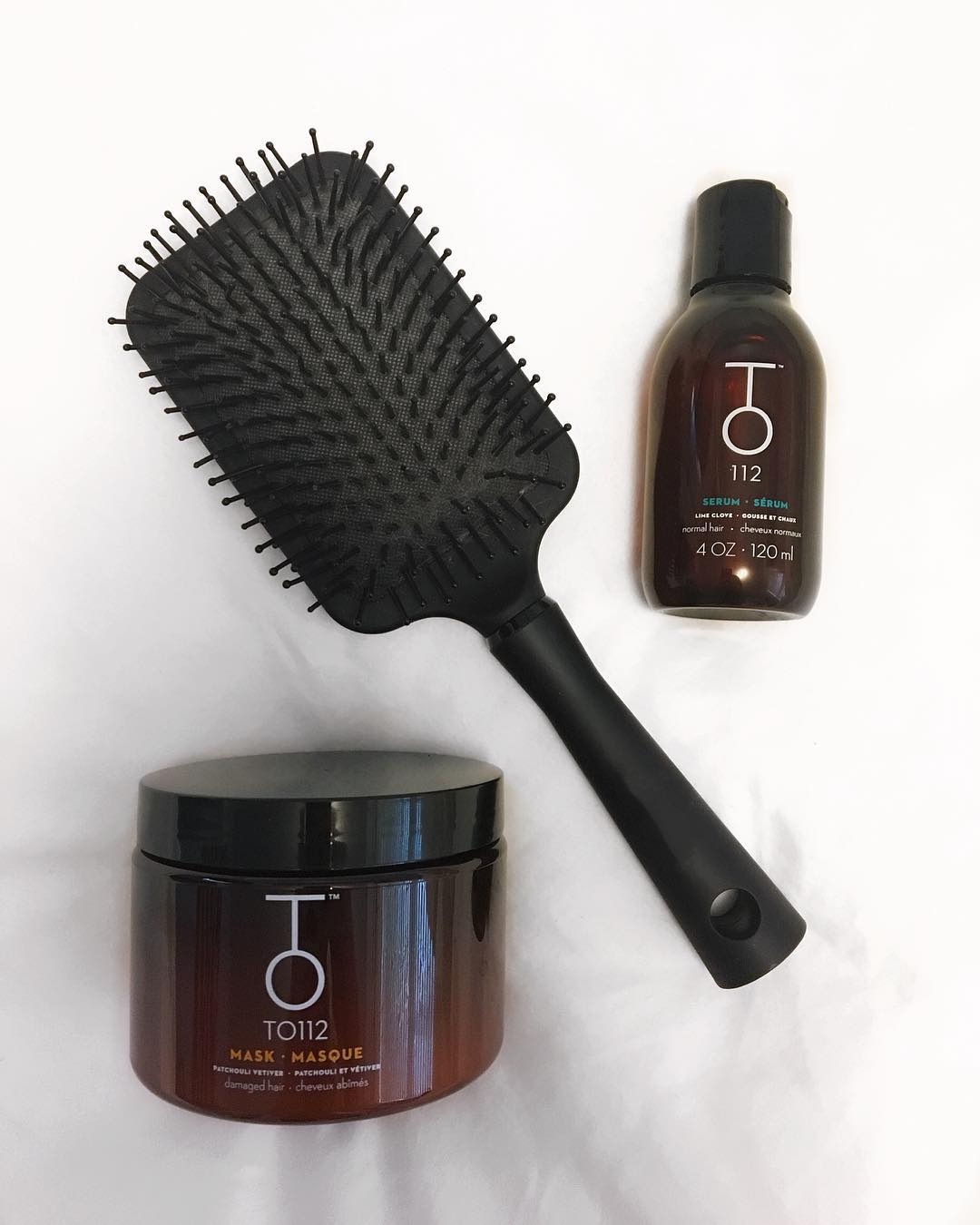 TO112 hair serum and to112 collagen mask on white bedsheet with black paddle brush facing up