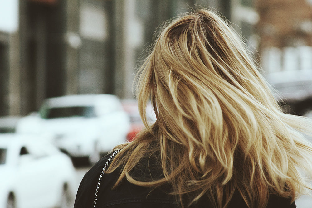 The back of a woman's head. She has medium length, fine, blonde hair. She is crossing a street.